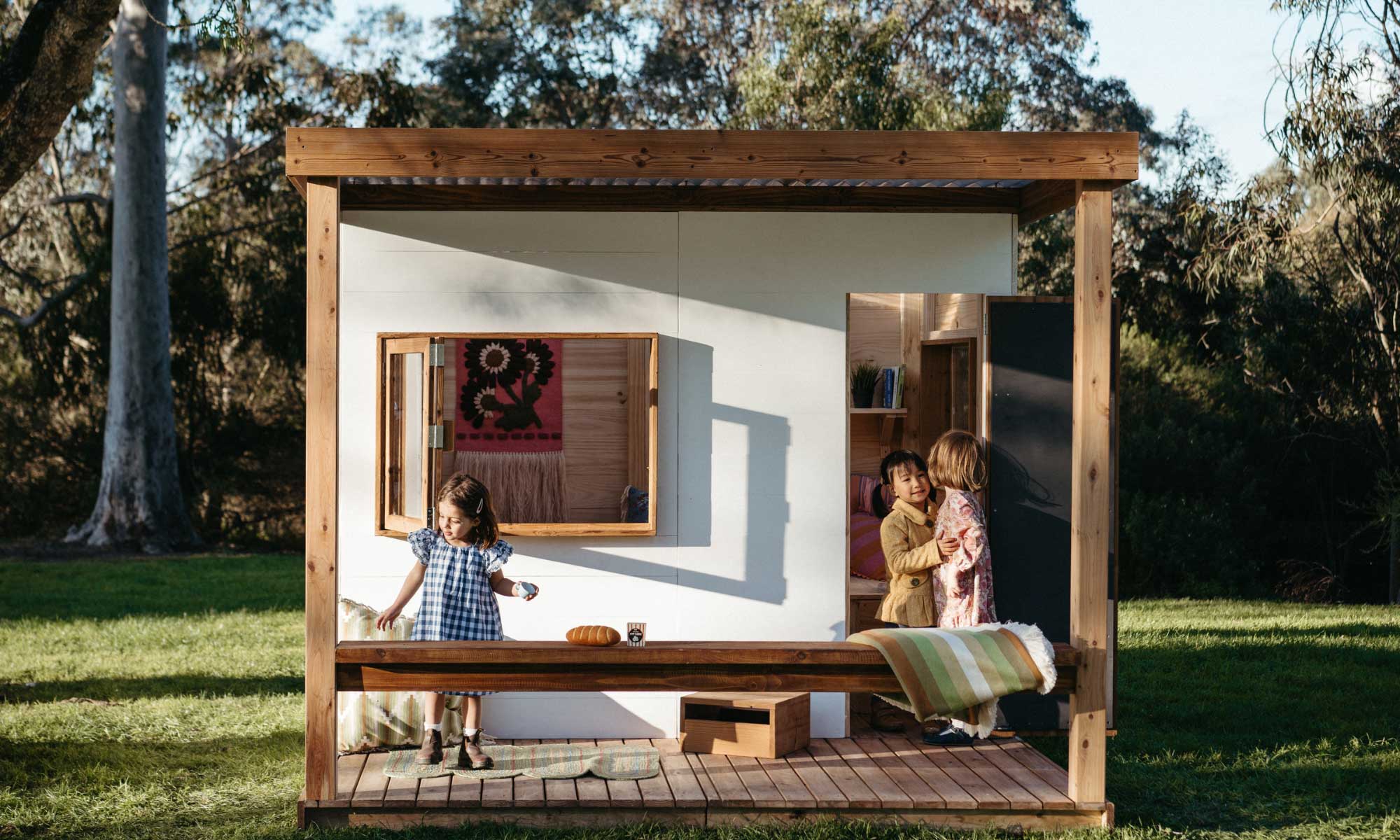 A Painted Wooden Cubby House in a natural bushland setting with 3 young girls playing in their cubby house. 