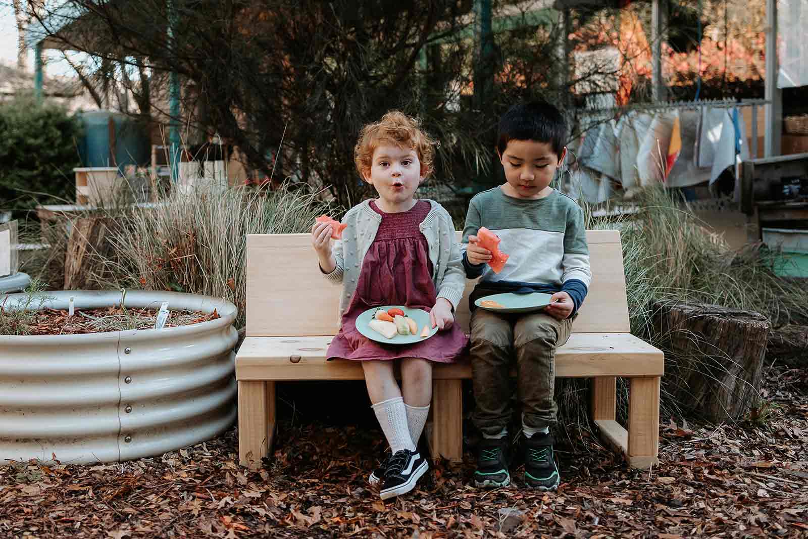 A picture of 2 young children in a childcare setting, sitting on a timber buddy bench eating watermelon. 